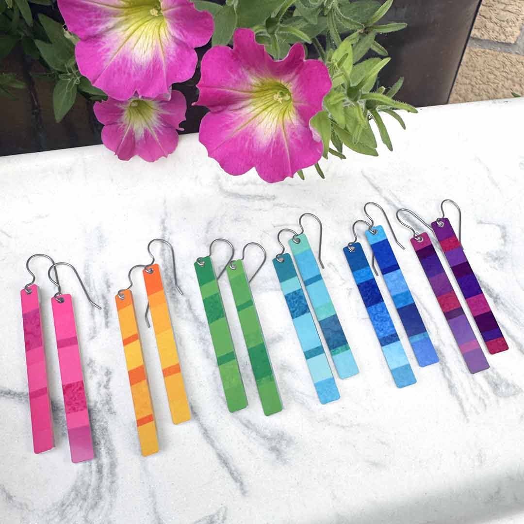 A set of colorful rectangular bar earrings with titanium fishhook wires, arranged in a rainbow gradient on a white marble surface with pink flowers in the background.