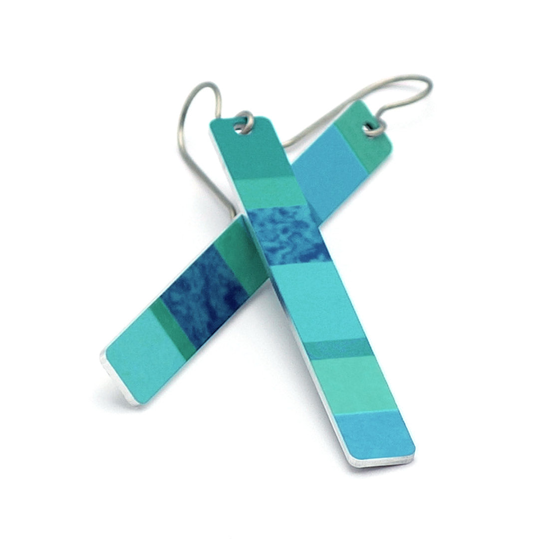 A pair of modern geometric dangle earrings featuring rectangular bar charms with a striped pattern of various shades of teal, blue, and green, suspended from titanium wire hooks on a white background.