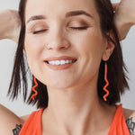 A close-up lifestyle shot of a woman with short dark hair, wearing bright orange wavy dangle earrings and an orange top against a light gray background.