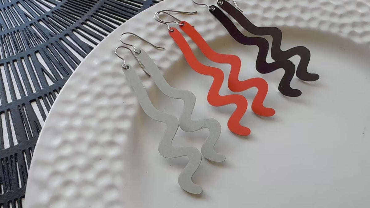 Video clip showing three long wavy pairs of dangle earrings in gray, orange, and black, displayed on a textured white plate.
