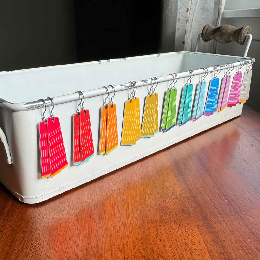 12 pairs of colorful quadrilateral earrings hanging from a metal tray on a wooden surface by ColorUpLife.