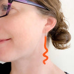 A close-up of a woman's ear wearing a bright orange wavy dangle earring, with part of her face, hair, and glasses visible against a plain light background.