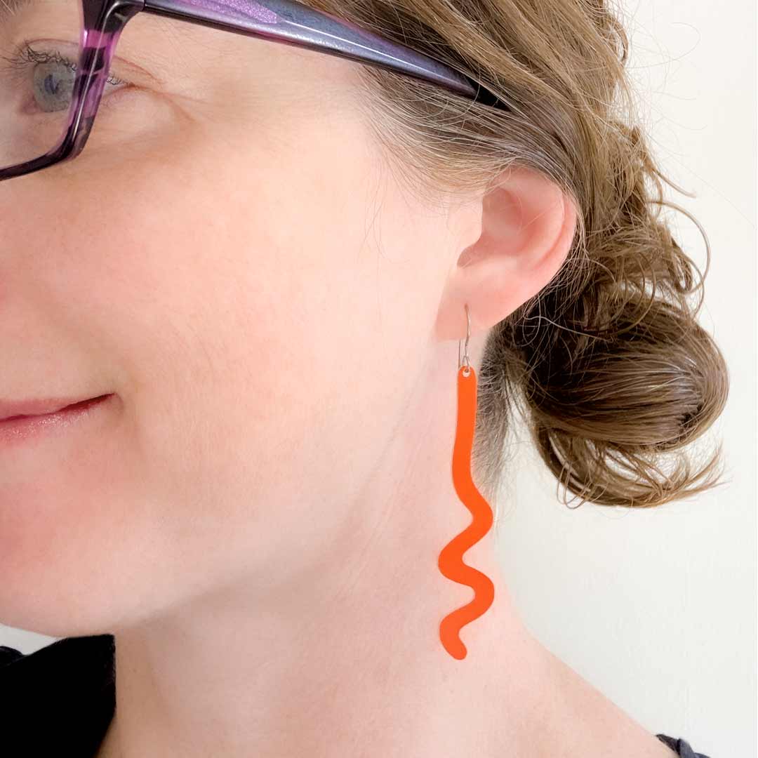 A close-up of a woman's ear wearing a bright orange wavy dangle earring, with part of her face, hair, and glasses visible against a plain light background.