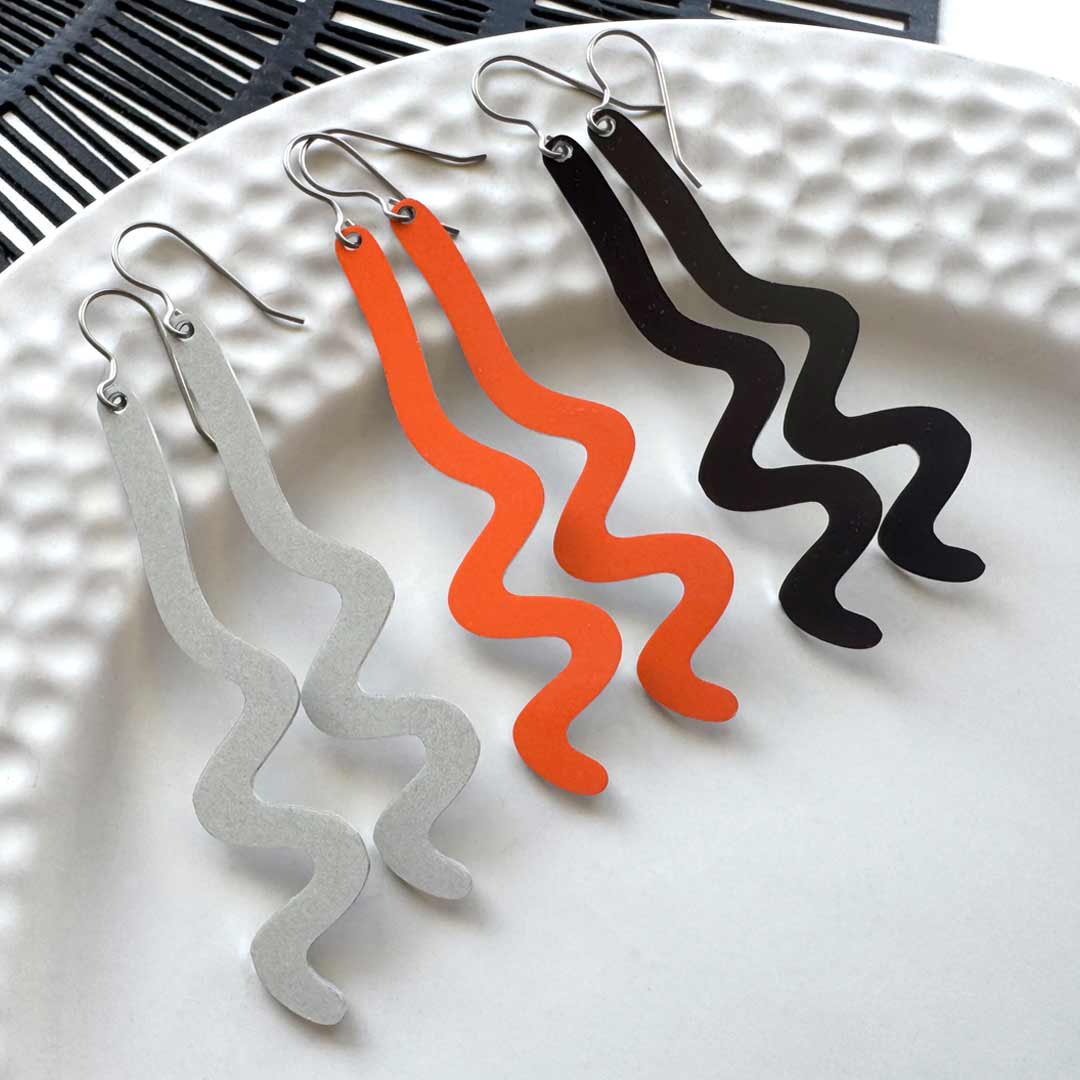 Three pairs of wavy dangle earrings in light gray, orange, and black, displayed on a white textured plate.